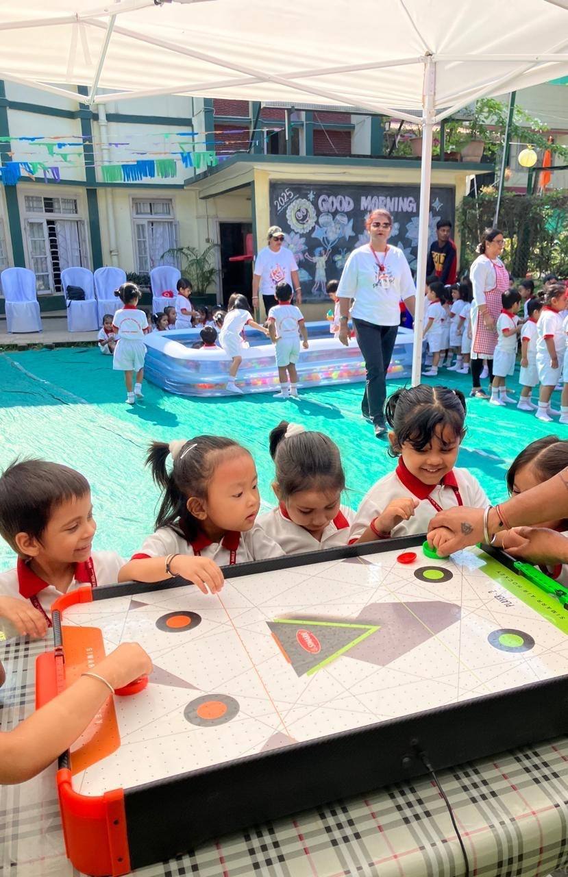Excited children in white and red uniforms play table air hockey under a white tent on the Good Shepherd Pre Primary School campus in Chandmari, Guwahati, while other children play in a ball pit and teachers watch near a “Good Morning 2025” chalkboard