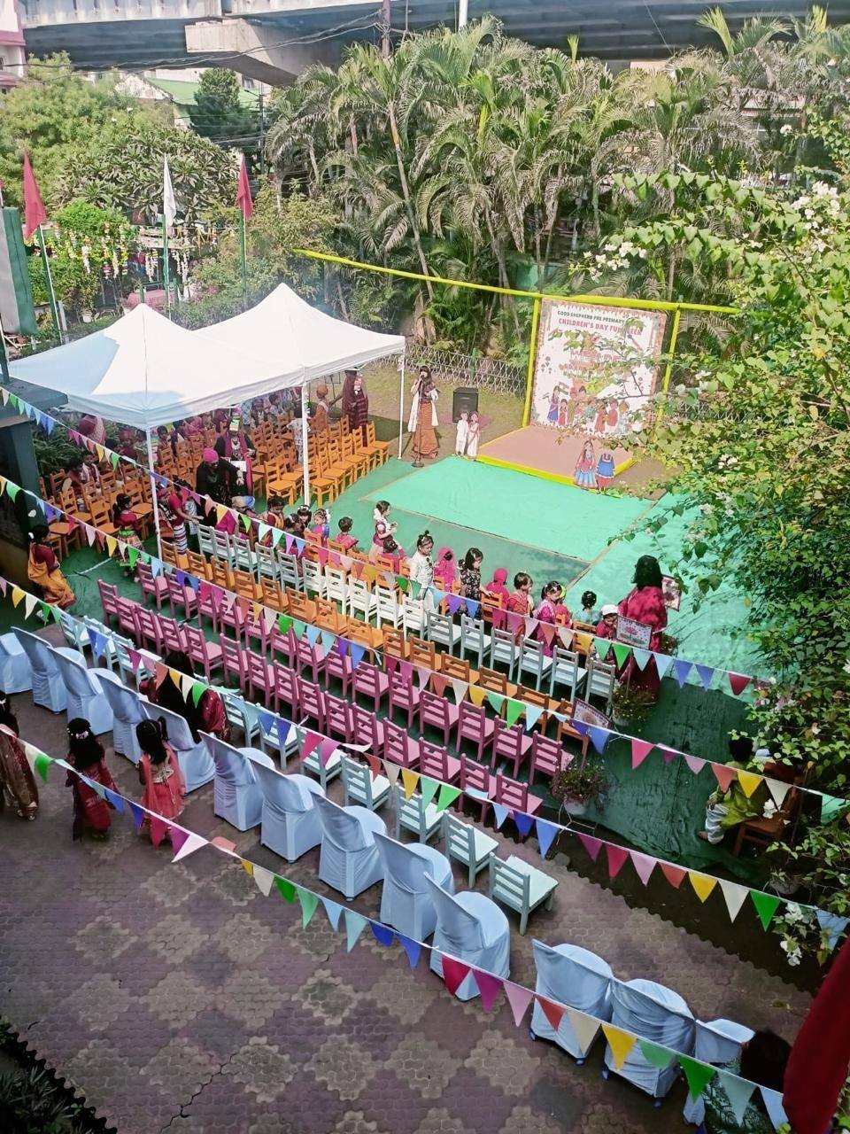 View from above of the school courtyard in Chandmari, Guwahati, set up for a Children’s Day function, with rows of chairs, colourful bunting, white tents, a stage with a banner and palm trees