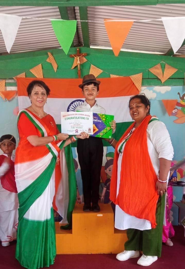 Two teachers in tricolour outfits present a certificate to a boy in a hat and moustache standing on a podium, with an Indian flag and tricolour bunting behind them at the school in Guwahati