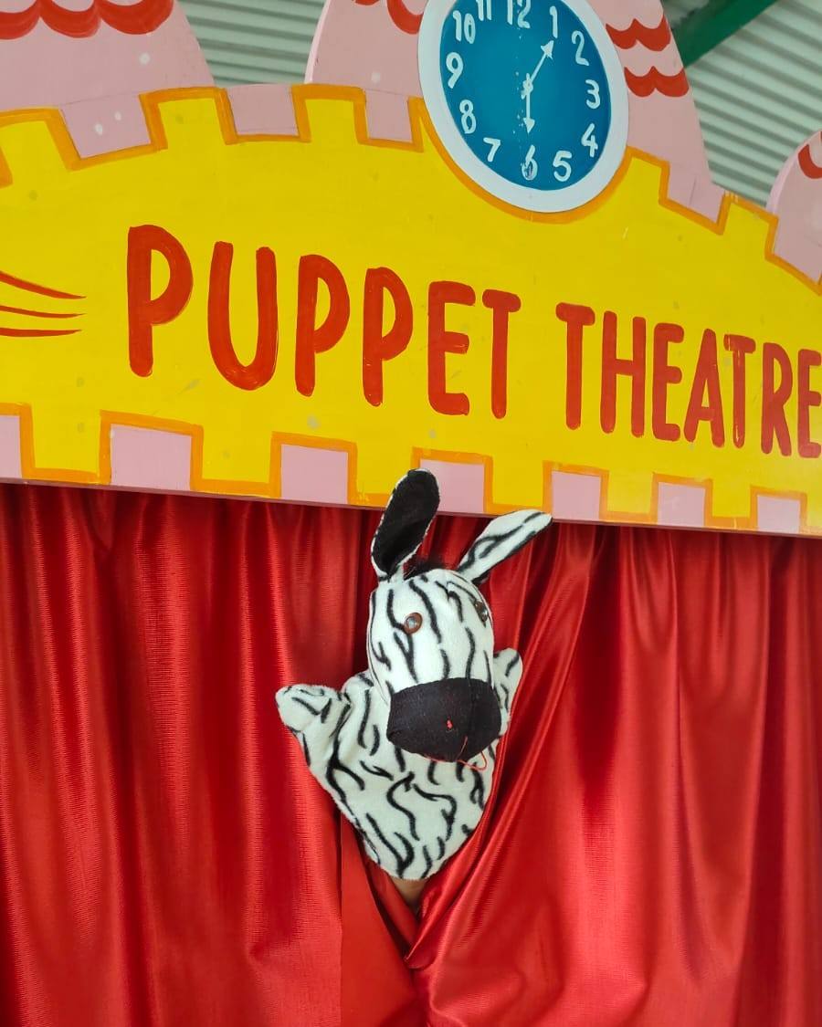 A zebra hand puppet peeks out between red curtains under a colourful “Puppet Theatre” sign at the school