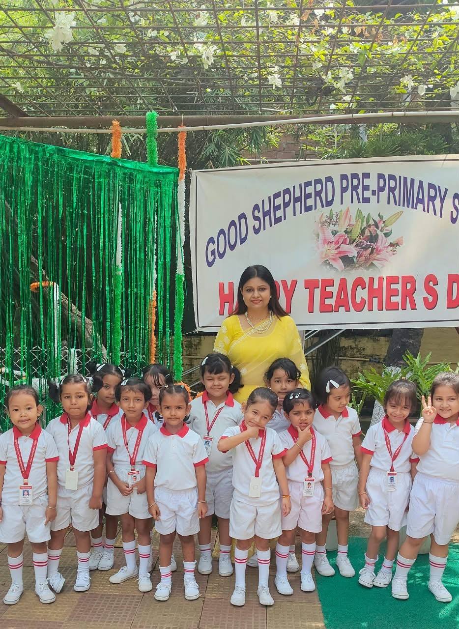 A teacher in a yellow saree stands with a group of smiling children in white uniforms beside a Good Shepherd Pre-Primary School Teachers’ Day banner